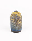 A small ceramic vase with a rounded body, featuring a speckled glaze that transitions from yellow and gray at the top to blue at the base, set against a plain white background.