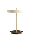 A modern table lamp with a circular off-white shade, a slim brass-colored metal stem, and a round dark bronze base, shown against a solid white background.