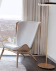 A modern lounge chair with a light gray upholstered seat and back, light wood frame, and angled legs, positioned next to a floor lamp in a bright room with a city view through a large window.