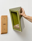 Hand showing a green ceramic bird house and the wooden attachment cleat on a white wall.