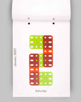 A white hanging calendar page showing the letter S formed by stacked rectangular blocks in lime green, orange, and reddish brown, with the text January 2027 and Saturday visible on a light gray background.