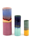 A set of three cylinder vases in different sizes with multicolored glazes. The largest vase is pink with blue and orange bands, the medium is green with a blue rim, and the smallest is predominantly blue with a yellow band.