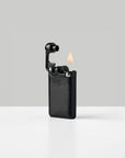 A black lighter with a textured metal body, shown open with a flame lit, set against a clean light gray background.