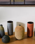 Six handmade ceramic vases in various shapes and earthy colors, including speckled gray, terracotta orange, textured beige, and dark green, arranged on a wooden shelf against a white wall.