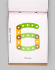 A top-down view of a hanging desk calendar page showing the number 6 in green and orange with the date December 2021 and the day Monday on a white page, set against a solid gray background.