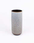 A tall, cylindrical ceramic vase with a speckled glaze that transitions from light gray at the top to a warm brown at the base, set against a plain white background.