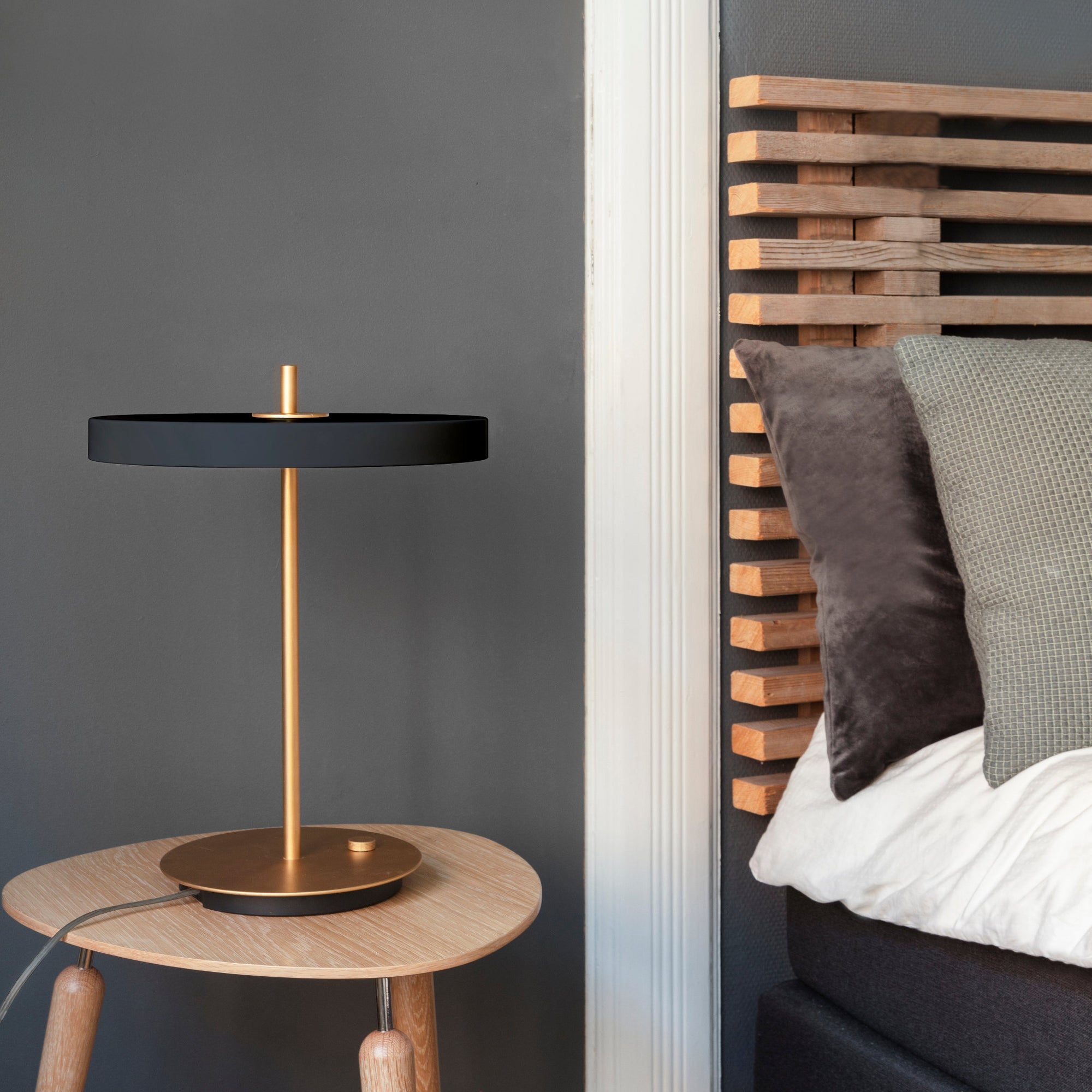 A modern table lamp with a matte black circular shade and a brushed brass gold base and stem, styled on a light wood side table next to a bed with a wooden headboard.