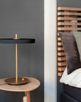 A modern table lamp with a matte black circular shade and a brushed brass gold base and stem, styled on a light wood side table next to a bed with a wooden headboard.