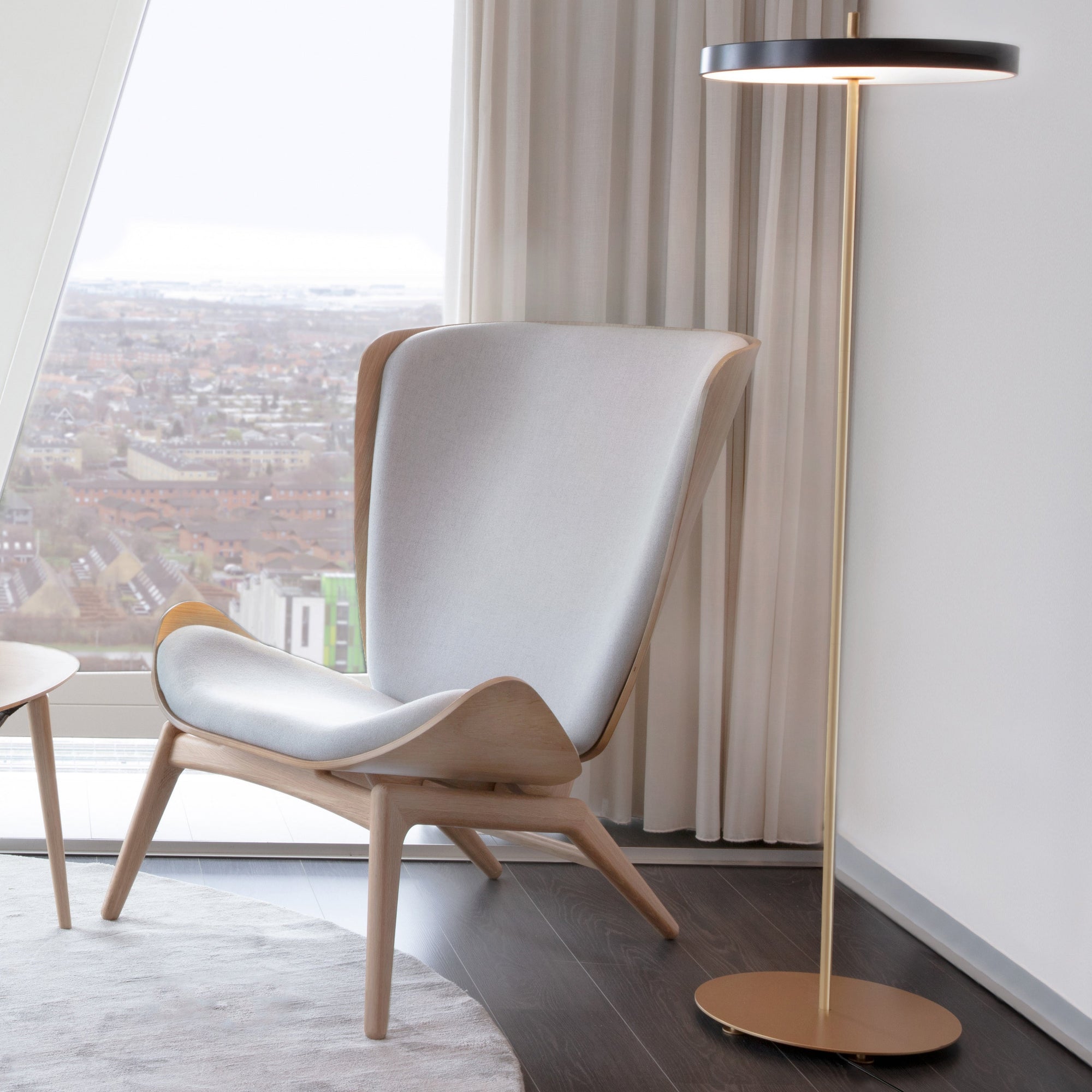 A modern lounge chair with a light gray upholstered seat and back, light wood frame, and angled legs, positioned next to a floor lamp in a bright room with a city view through a large window.