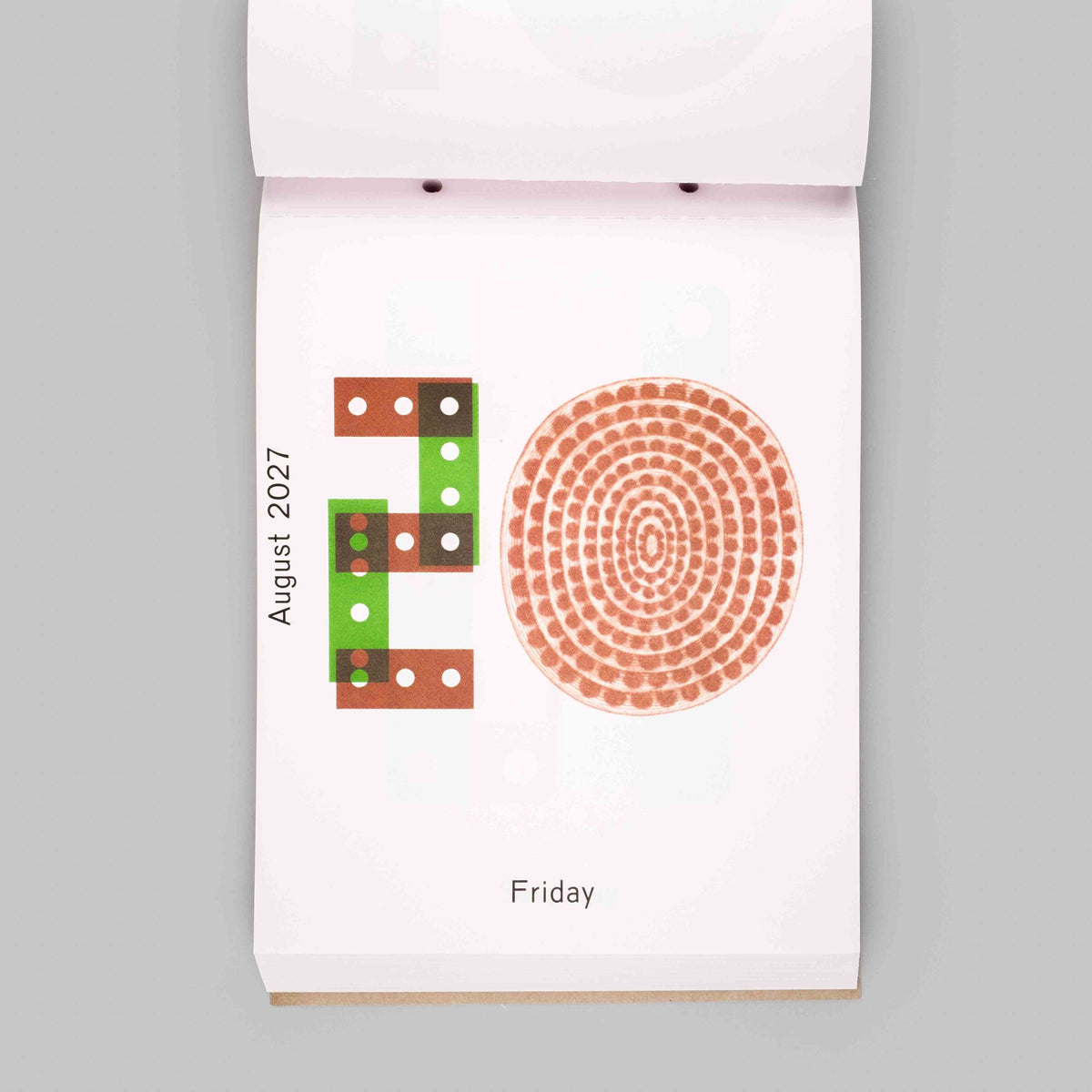 A single-page calendar page from a hanging wall calendar, showing the day Friday with a decorative orange dot pattern and a green-and-brown block design, photographed against a plain light gray background.