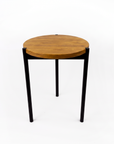 A modern round side table with a warm medium-brown wood top and slim black metal legs, photographed against a seamless white background.