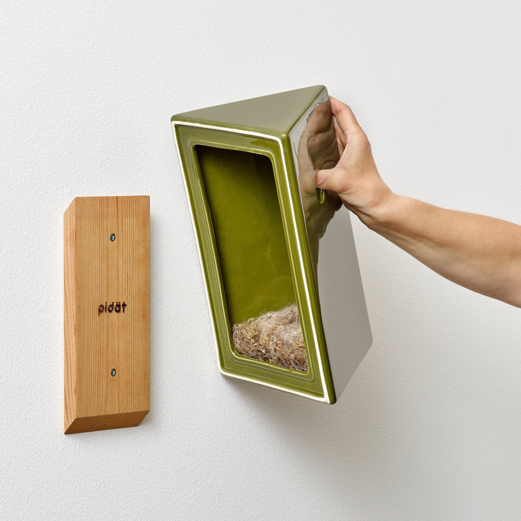 Hand showing a green ceramic bird house and the wooden attachment cleat  on a white wall.