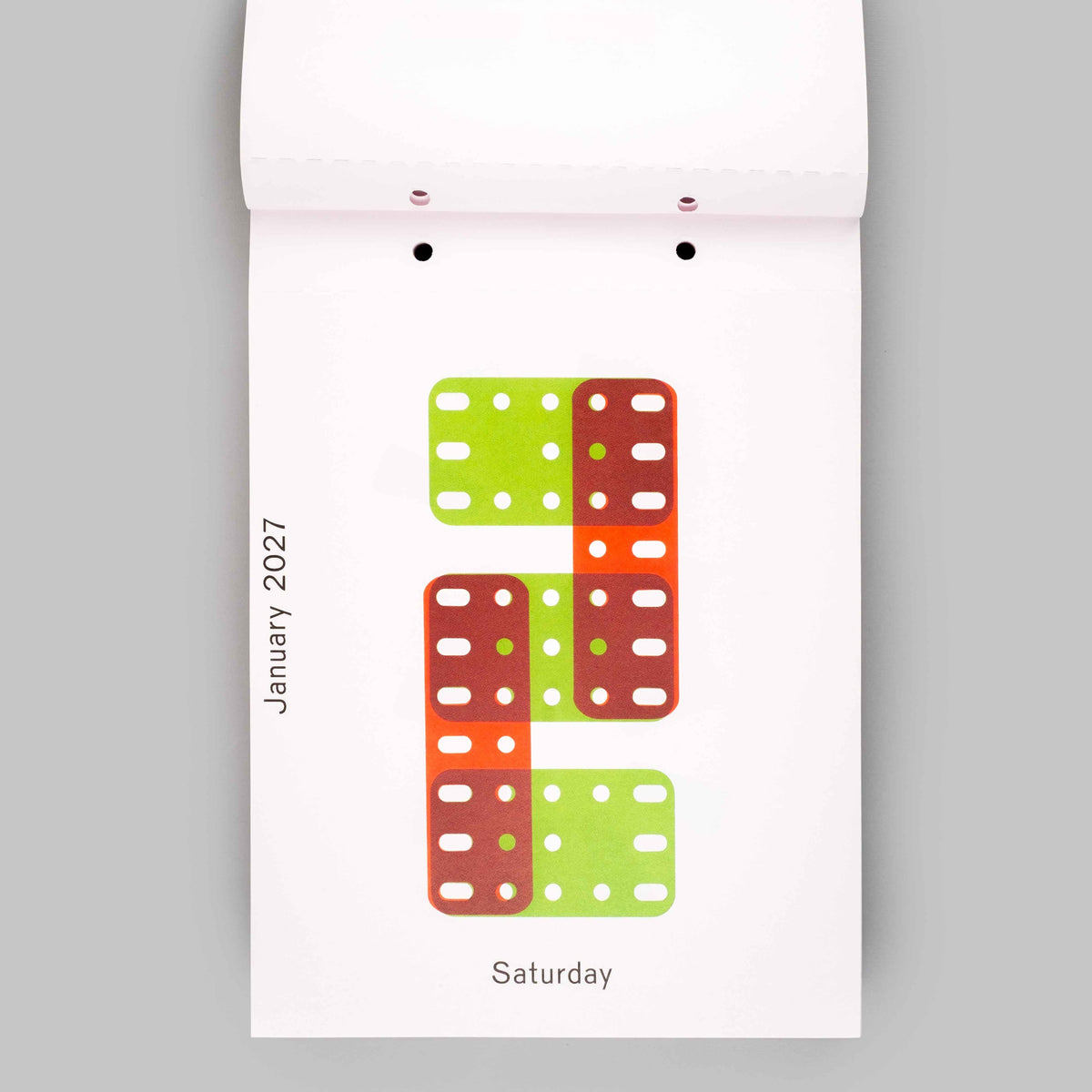 A white hanging calendar page showing the letter S formed by stacked rectangular blocks in lime green, orange, and reddish brown, with the text January 2027 and Saturday visible on a light gray background.
