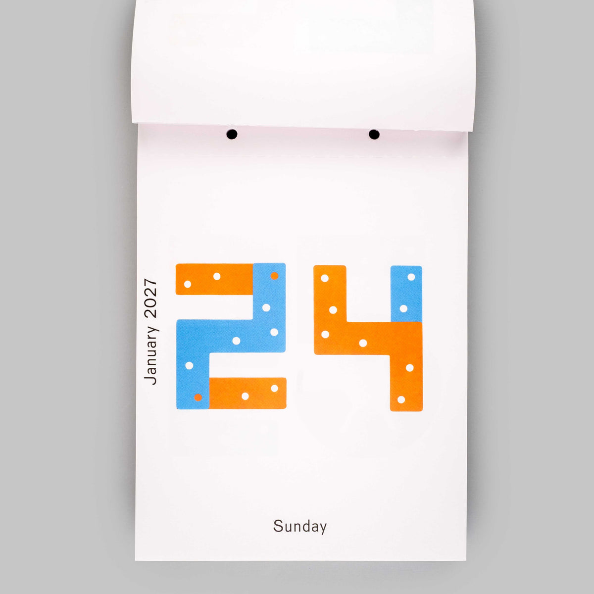 A minimalist white calendar page showing the number 24 in blue and orange with small white dots, labeled January 2021 and Sunday, photographed against a light gray background.