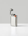 A brushed stainless steel lighter with a small orange tipped swing arm, standing upright on a white surface against a light gray background.