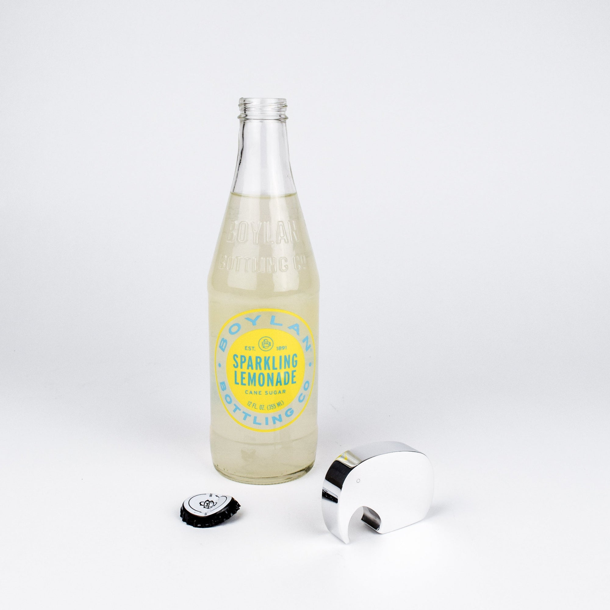 A clear glass bottle of Boylan Sparkling Lemonade with a yellow label stands on a white studio background, alongside a silver bottle opener and a black bottle cap.