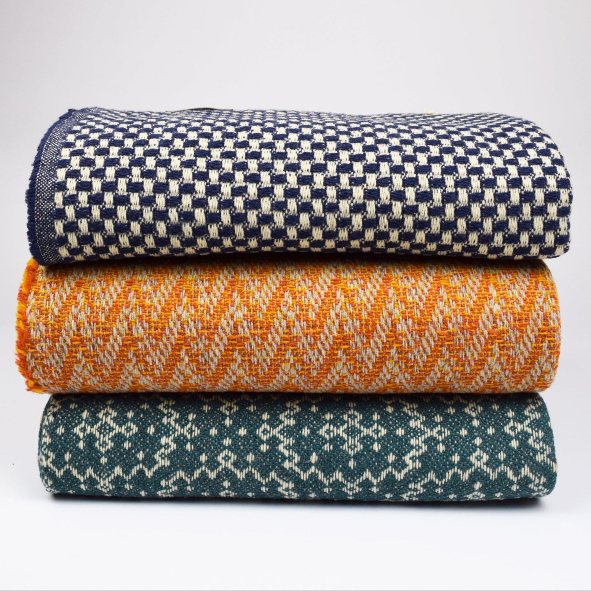 Stack of three woven blankets in navy, orange, and teal on a light gray background