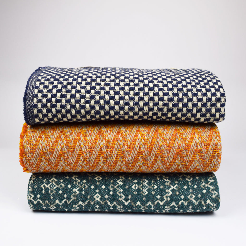 Stack of three woven blankets in navy, orange, and teal on a light gray background