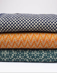 Stack of three woven blankets in navy, orange, and teal on a light gray background