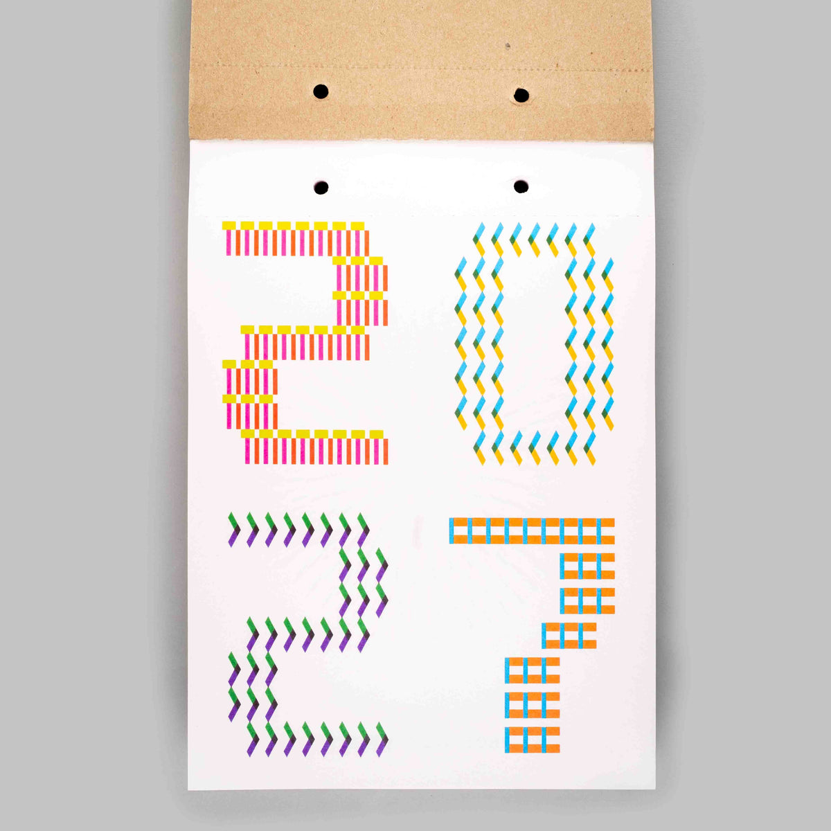 A white rectangular card or print with colorful geometric numbers and symbols arranged to spell '2027', attached to a brown kraft paper backing with two small black dots near the top, shown against a light gray background.