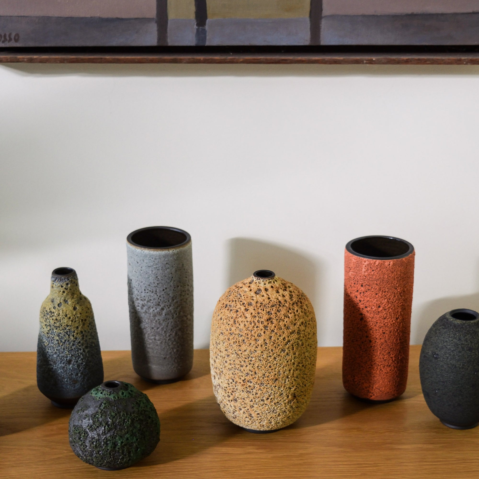 Six handmade ceramic vases in various shapes and earthy colors, including speckled gray, terracotta orange, textured beige, and dark green, arranged on a wooden shelf against a white wall.
