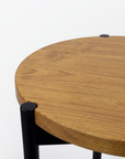 Close-up of a round wooden table top with a warm medium-brown wood grain finish and black metal legs, shown against a plain white background.
