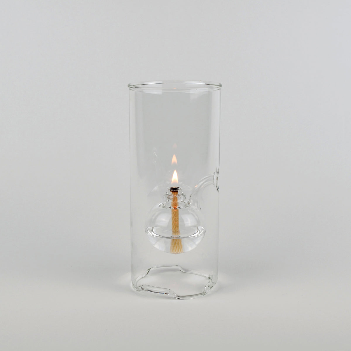 A clear glass cylindrical holder with a small glass oil lamp inside, lit with a visible flame, photographed against a plain light gray studio background.