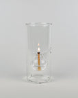 A clear glass cylindrical holder with a small glass oil lamp inside, lit with a visible flame, photographed against a plain light gray studio background.