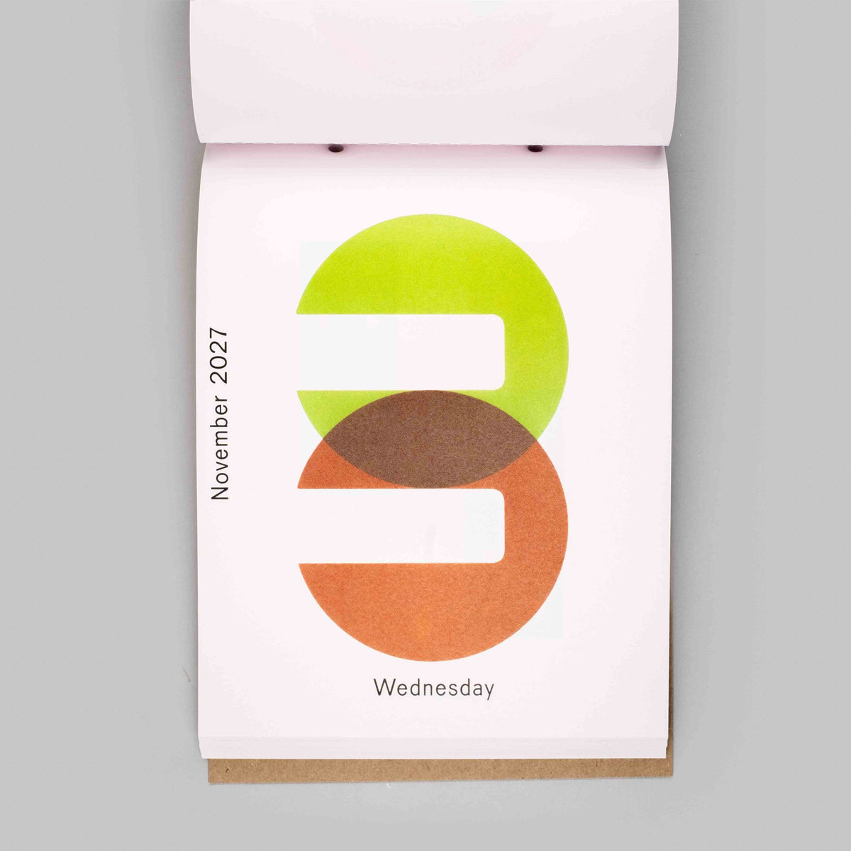 A single-page desk calendar or planner page shown open against a light gray background. The page is white and features a large overlapping green and orange graphic with the text 'November 2027' vertically on the left and 'Wednesday' at the bottom.