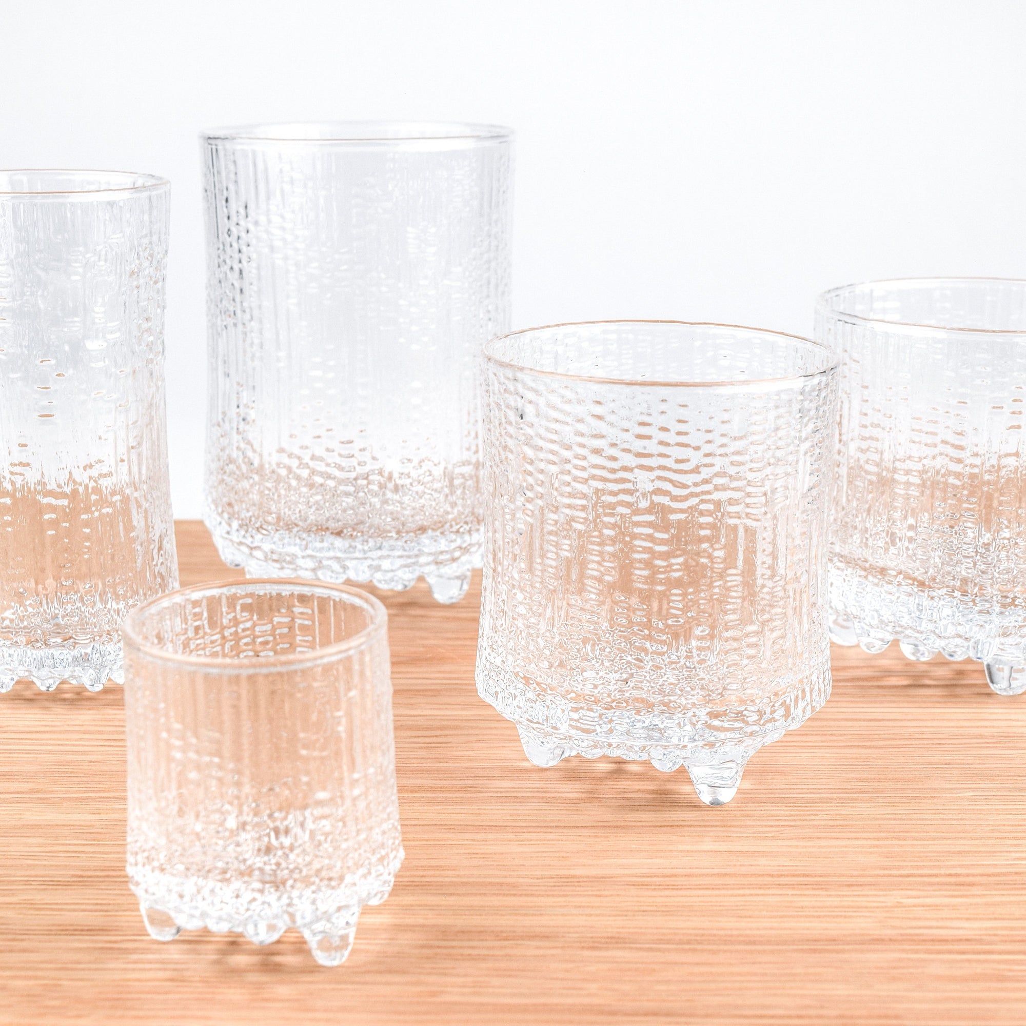Five clear textured glass tumblers with footed bases arranged on a light wood surface against a white background.