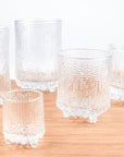 Five clear textured glass tumblers with footed bases arranged on a light wood surface against a white background.