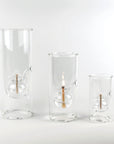 Three clear glass candle holders in graduated sizes, each featuring an internal glass bulb with a wick and a lit flame, set against a seamless white background.