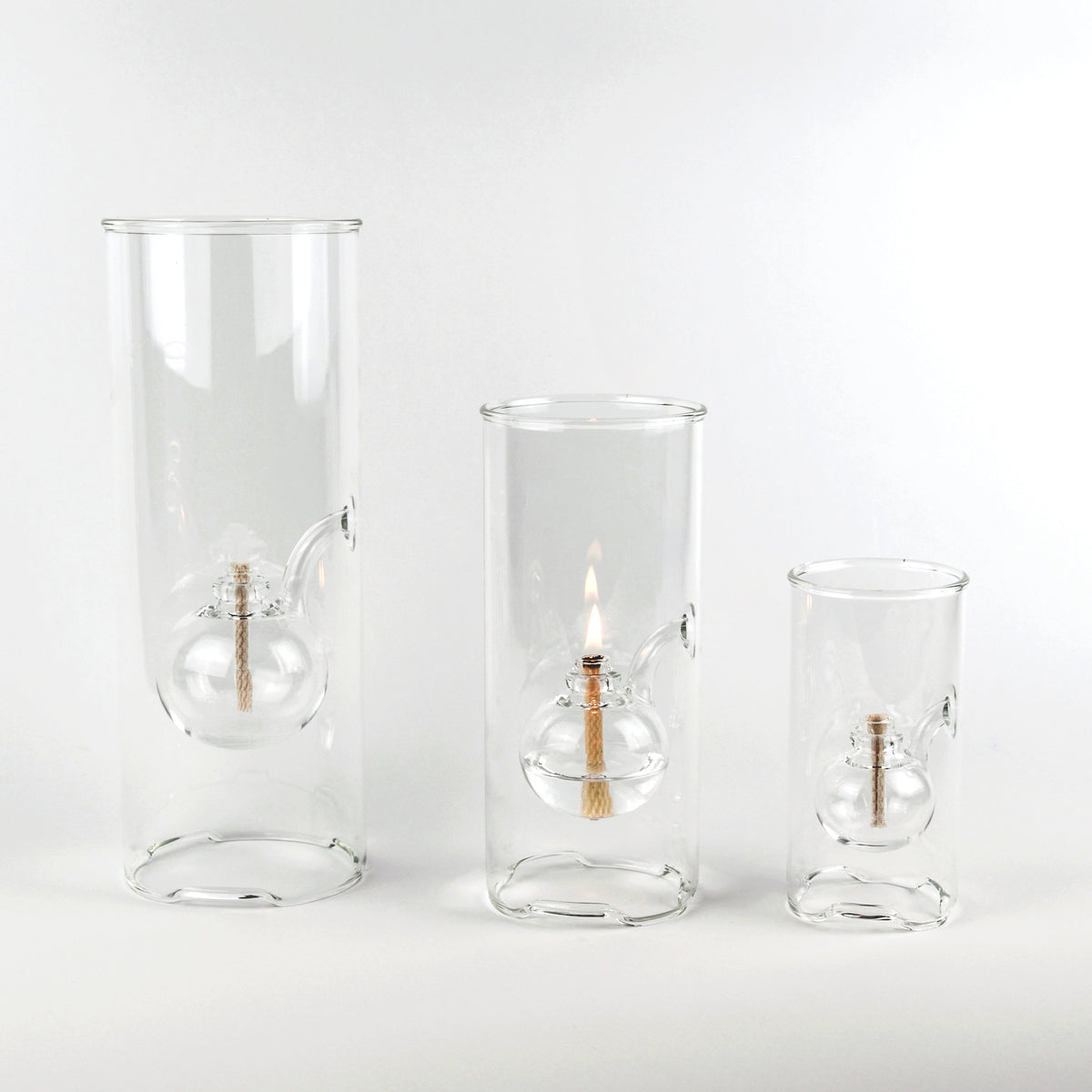 Three clear glass candle holders in graduated sizes, each featuring an internal glass bulb with a wick and a lit flame, set against a seamless white background.